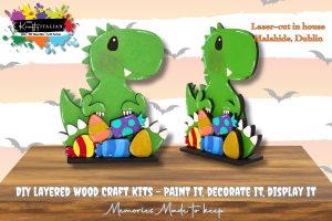 Two green dinosaur-shaped wooden craft kits, painted and decorated, stand on a surface with colourful eggs at their feet. Text describes them as DIY layered wood craft kits.