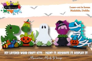 Four colourful Halloween-themed wooden craft figures—a dinosaur, a ghost, a witch, and a dragon—are displayed on a wooden surface with a bat and cloud background.