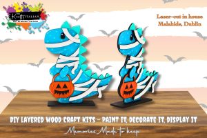 Two blue dinosaur wood craft figures dressed as mummies, holding orange pumpkin buckets, displayed on stands. Background text promotes DIY craft kits from Malahide, Dublin.