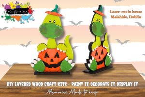 Two painted, layered wood craft dinosaurs holding jack-o’-lanterns, displayed on stands with bats in the background. Text advertises DIY craft kits and laser cutting in Malahide, Dublin.