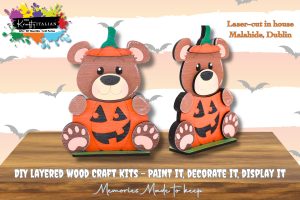 Two wooden bear figures wearing pumpkin costumes stand on green bases. The text promotes DIY layered wood craft kits for painting and display.