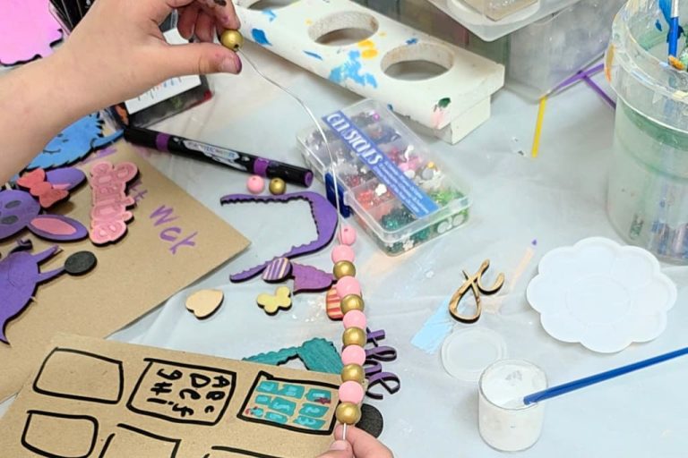 Two hands thread colourful beads onto a wire at a crafting table covered with art supplies, including cut-out shapes, paint, brushes, a marker, and a palette—an engaging example of fine motor skills activities for children.
