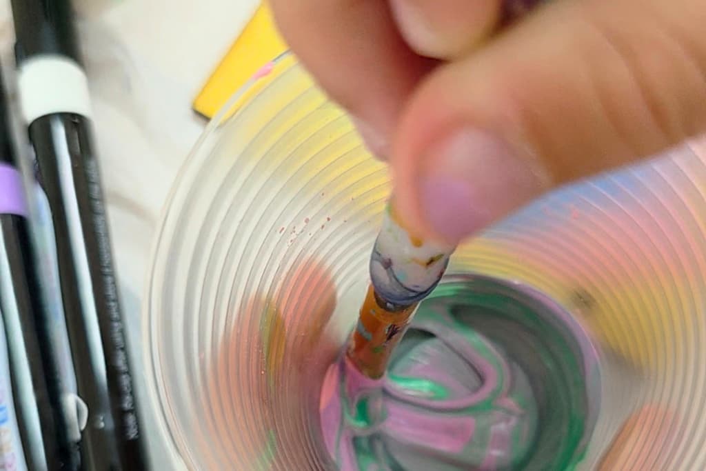 A hand stirs a paintbrush in a plastic cup filled with swirled green and pink paint. Nearby, there are black and white markers, highlighting the benefits of crafting for children as they explore creativity through colours and art tools.