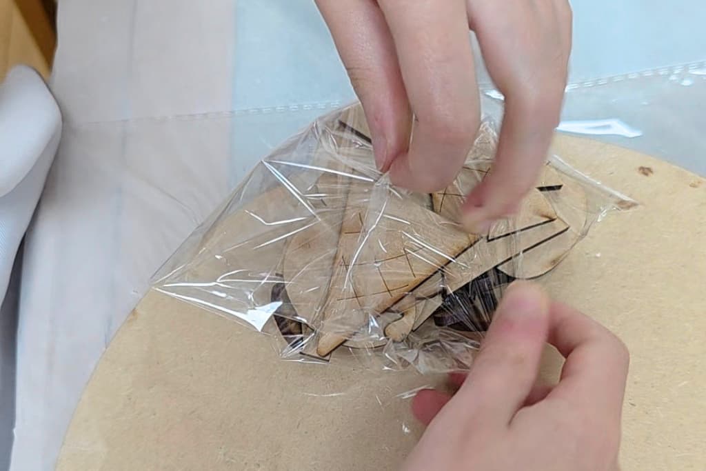 Two hands carefully unwrap a small bundle of wooden pieces from a sheet of clear plastic film on a light-coloured surface, illustrating the benefits of crafting for children as they develop fine motor skills and creativity through hands-on activities.