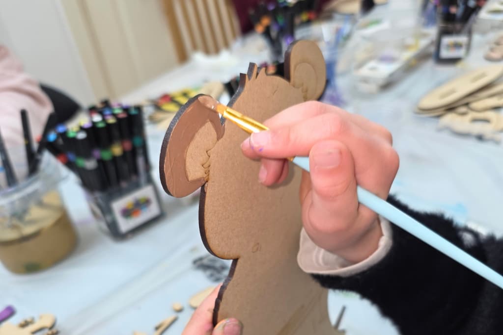 A person paints a brown ear on a cardboard animal cut-out with a small brush, demonstrating the benefits of crafting for children; art supplies and felt-tip pens are visible on the table in the background.