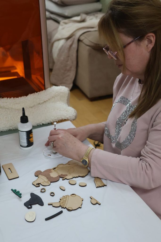 A woman assembling wooden craft pieces at a table with glue, various cut-outs, and tools in front of her—capturing the creative spirit that's all about The Krafty Italian.