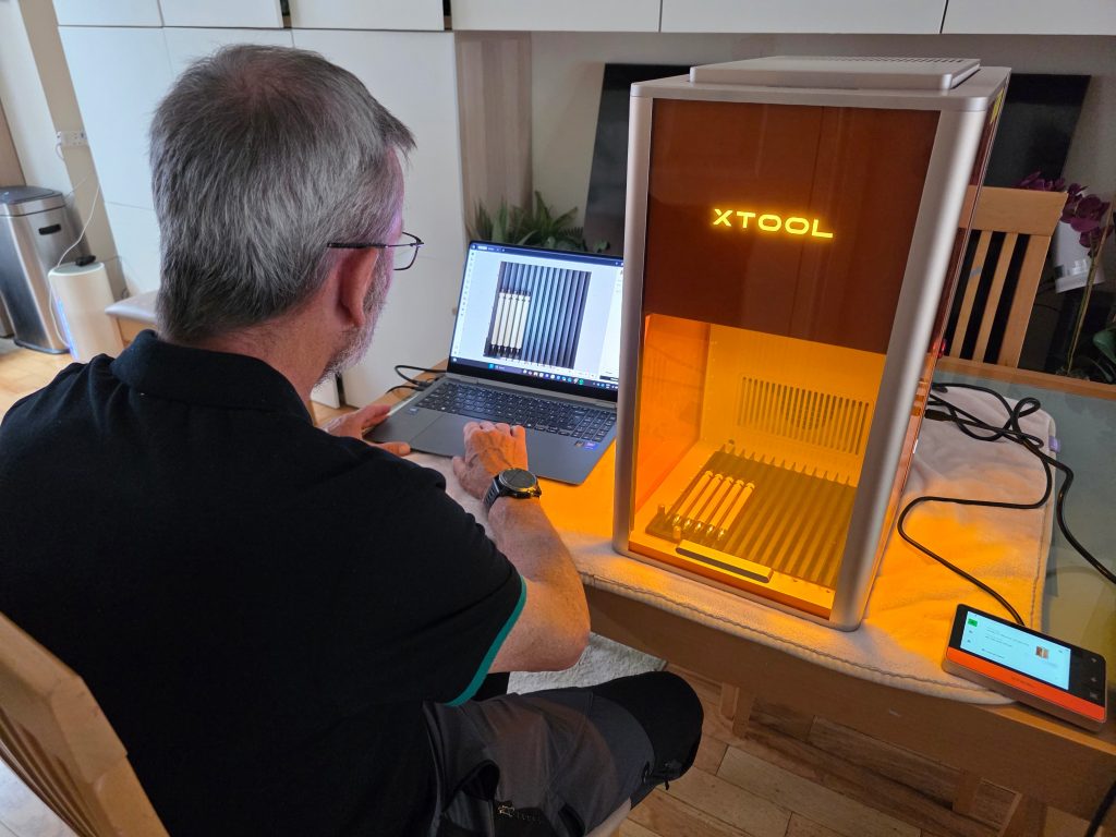 A person operates a laptop connected to an xTool laser engraving machine, which is active on a table beside them—perhaps working on a project about the Krafty Italian.