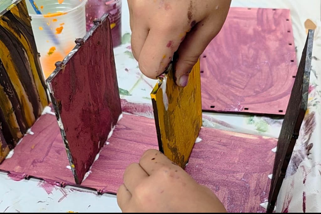 A child’s hands assemble and glue painted wooden pieces, building a small colourful structure on a paint-covered table, highlighting the benefits of crafting for children such as creativity and fine motor skill development.