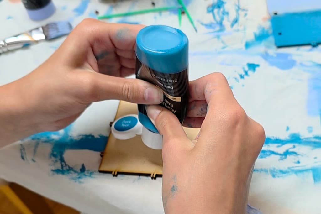 Two hands holding a bottle of blue acrylic paint, squeezing paint onto a small round palette. The table is covered with a white cloth stained with blue paint, and art supplies are nearby—a colourful scene that highlights the benefits of crafting for children, such as fostering creativity and enhancing fine motor skills.