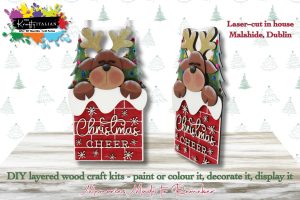 Two Christmas Cheer Reindeer Hanging Sign DIY kits featuring wooden reindeer decorations with festive signs, snowy accents, and decorated Christmas trees in the background. Promotional text highlights a variety of DIY craft kits—including a Halloween sweet basket shelf sitter kit—and local laser cutting services in Malahide, Dublin.