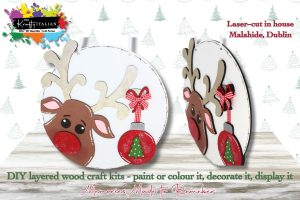 Two round, white Reindeer & Bauble Round Hanging Sign DIY kits are displayed, featuring wooden reindeer shapes with antlers, festive red bows, and Christmas baubles. The text notes they are laser-cut in Malahide, Dublin and ready for you to paint and decorate—perfect as a festive shelf sitter or to spark ideas for your next Halloween sweet basket DIY kit. The background features subtle holiday designs.