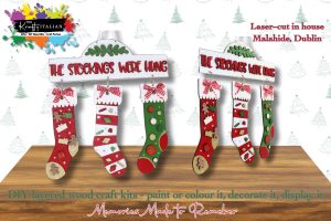 Two “The Stockings Were Hung Hanging Sign” DIY kits hang from labelled mantel signs, creating a charming shelf sitter display. Each kit includes three festive stockings ready to be decorated. The text highlights that they’re laser-cut in Malahide, Dublin—ideal for painting or colouring. The background features subtle tree graphics, bringing seasonal cheer reminiscent of Halloween sweet basket fun.